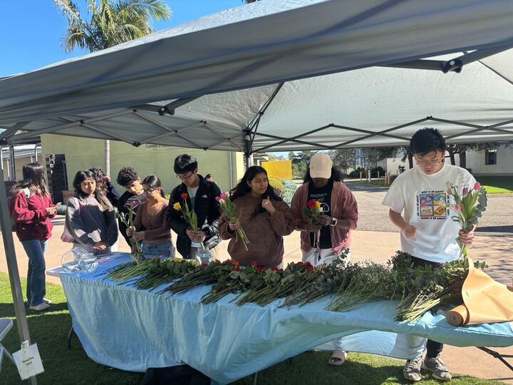 9 students stand in line in front of a table beneath a tent. The table has many flowers sitting on it in organized piles, and most students are creating bouquets with the flowers.